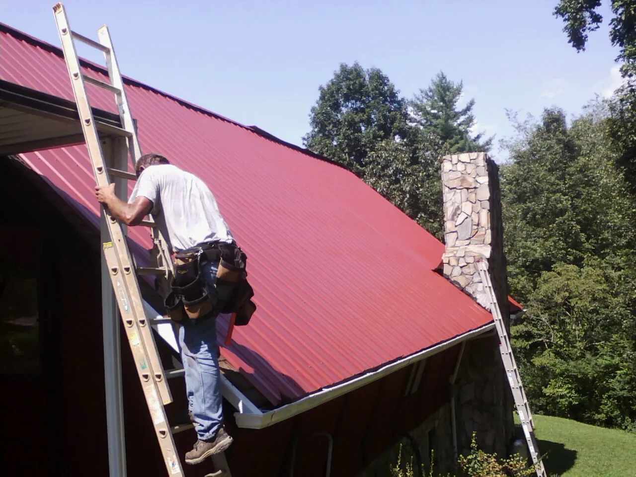 Roofer on a ladder beside a red metal roof and stone chimney — Soffit & Fascia Repair by Roofing Brothers in North Apollo
